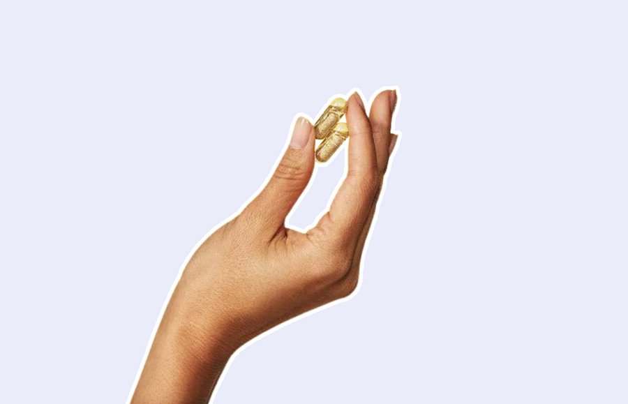 Hand held up against a purple background, with 2 vitamin capsules in between fingers. Hand prompts answer to what vitamin deficiency causes hair loss and what is needed in her diet to stop hair loss.
