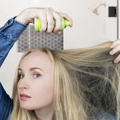 woman using a diy dry shampoo on her hair in front of a mirror