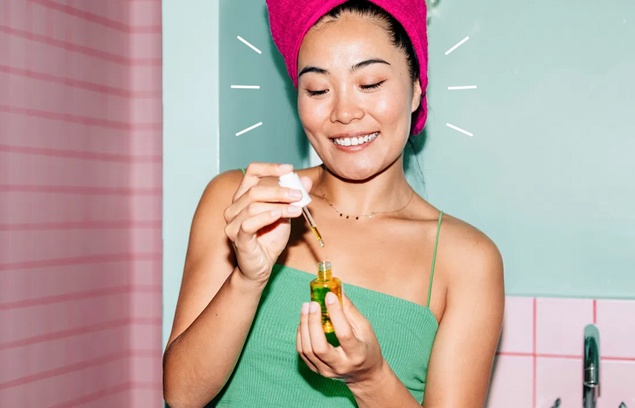 Asian woman photographed with pink hair towel following a glowing up guide; bottle of facial oil in hand