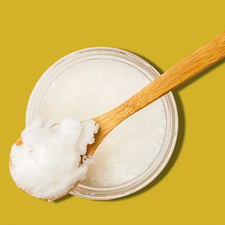 cup of coconut oil on yellow background; model off frame using benefits of coconut oil on skin